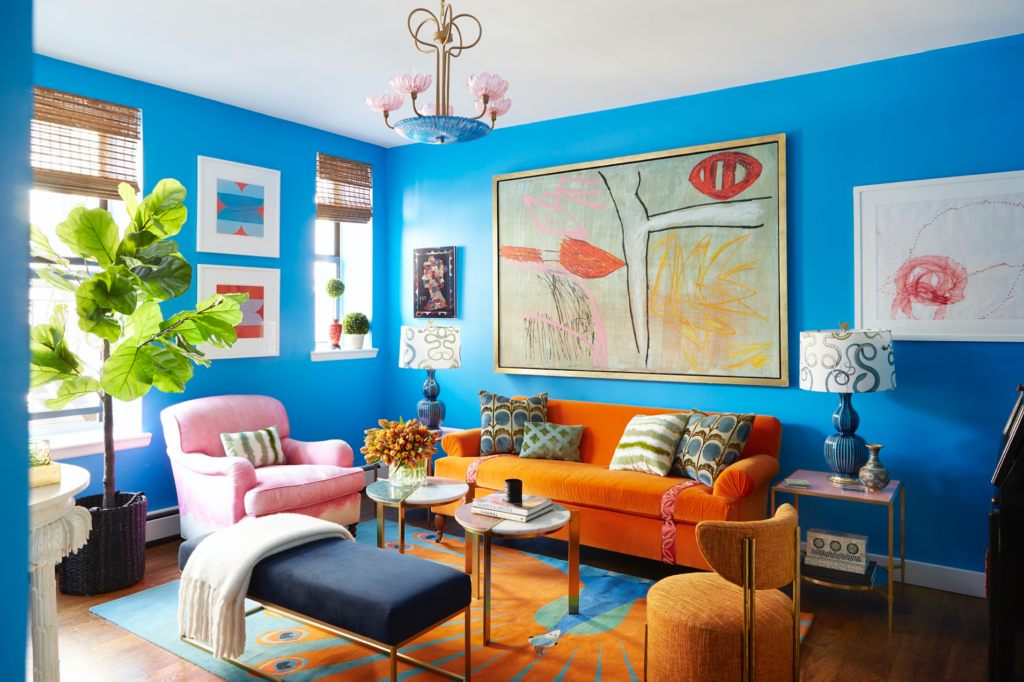 Living room with vibrant walls and abstract art frames on them, orange and pink couches with colorful cushions on an orange carpet, with a plant by the window